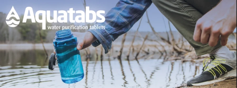 Person crouched at a lakeshore filling a water bottle with Aquatabs water purification tablets