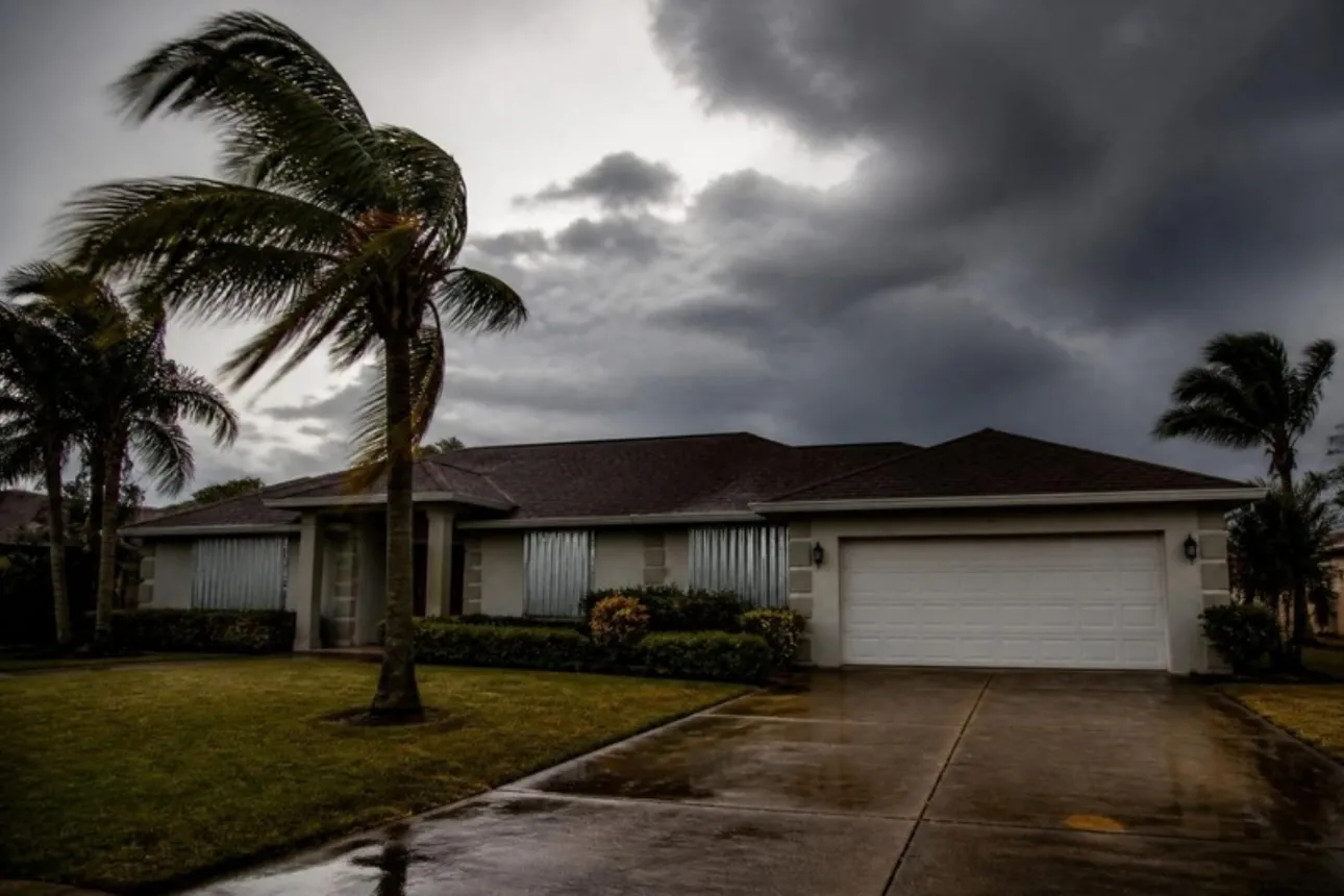 Single-story Florida home with hurricane shutters closed as palm trees bend under dark storm clouds