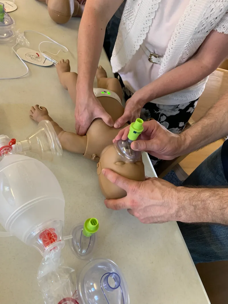 Students practicing infant CPR on a training manikin, one giving chest compressions while another seals a rescue breathing mask