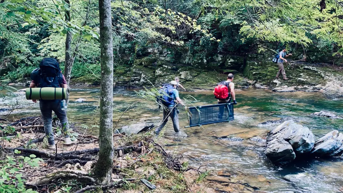 Wilderness first aid students carrying a rescue litter across a shallow forest river with backpacks on